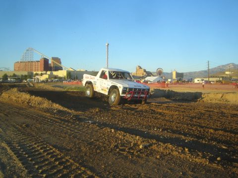 1990 Ford Bronco - white off-road race truck with a tube front bumper rounds a banked dirt corner, with an amusement park and mountains behind it.
