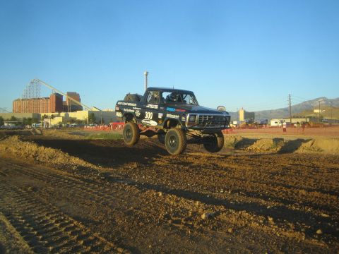 1990 Ford Bronco - black off-road race truck marked 300 catches air over a dirt course, with a roller coaster and mountains in the background.