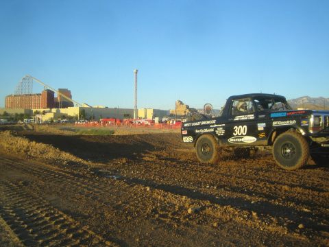 1990 Ford Bronco - black off-road race truck lettered West Coast Broncos and number 300 sits on a dirt short-course track near an amusement park.