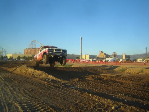 1990 Ford Bronco - red-and-white off-road race truck catches air off a dirt jump on a short-course track, with an amusement park behind.