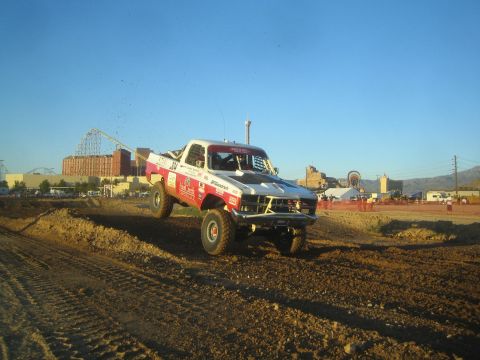 1990 Ford Bronco - white off-road race truck with a hood scoop catches air over a dirt course, with a roller coaster and mountains in the background.