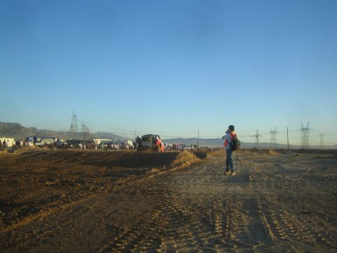 1990 Ford Bronco - person wearing a backpack stands on a dirt course at golden hour watching distant race vehicles under power lines and hills.