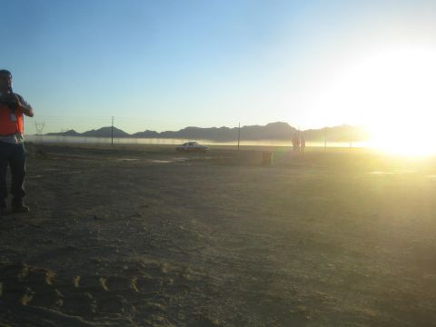 1990 Ford Bronco - man in an orange safety vest stands on a dusty desert lot as a car is silhouetted against the low sun and distant mountains.