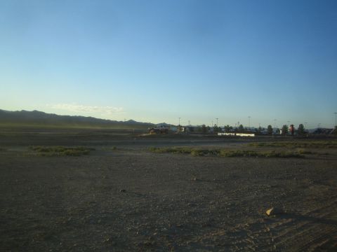 1990 Ford Bronco - wide expanse of open dirt desert lot with sparse scrub, distant buildings, and a mountain ridge under a clear blue sky.