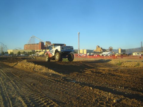 1990 Ford Bronco - blue-and-white off-road race truck launches off a dirt jump on a short-course track, with an amusement park skyline behind.