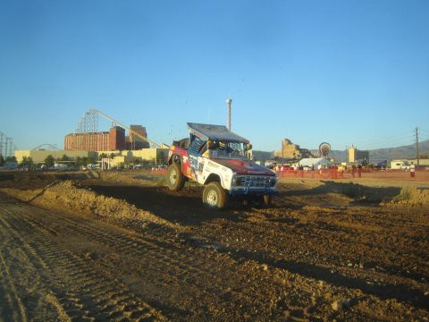 1990 Ford Bronco - colorful off-road race truck with its hood raised sits on a dirt short-course berm, with an amusement park and crowd behind.