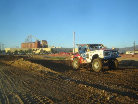 1990 Ford Bronco - red-white-and-blue off-road race truck climbs a dirt track berm at golden hour, with a roller coaster and crowd behind.