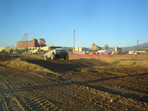 1990 Ford Bronco - light-colored off-road truck sits on a dirt track berm at golden hour, with a roller coaster and amusement park buildings behind.