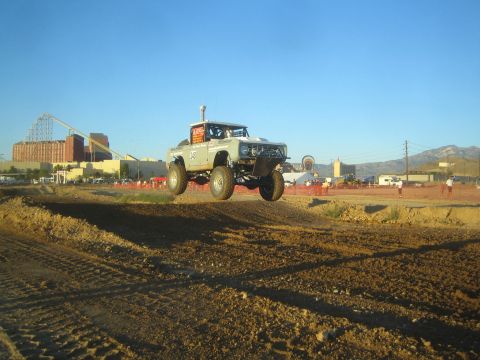 1990 Ford Bronco - white off-road race truck catches air off a dirt jump on a short-course track, with an amusement park skyline and mountains behind.
