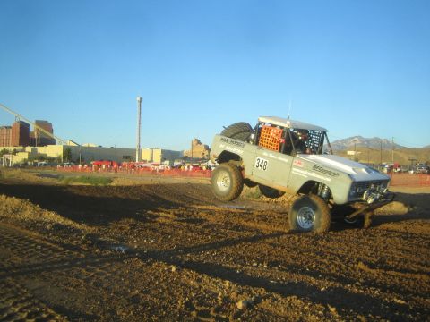 1990 Ford Bronco - white early Ford Bronco-style race truck marked 348 launches off a dirt berm at an off-road racing event near a casino-style building.