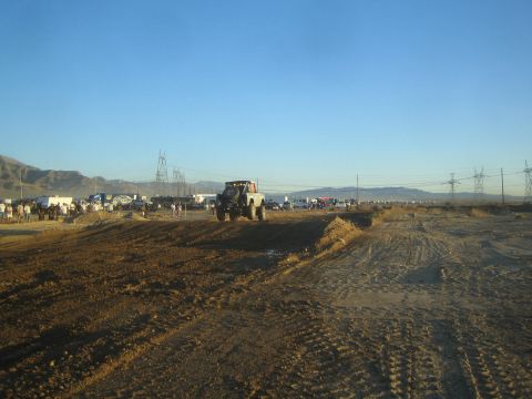 1990 Ford Bronco - off-road race truck goes airborne over a dirt jump on a desert course lined with transmission towers and distant spectators.