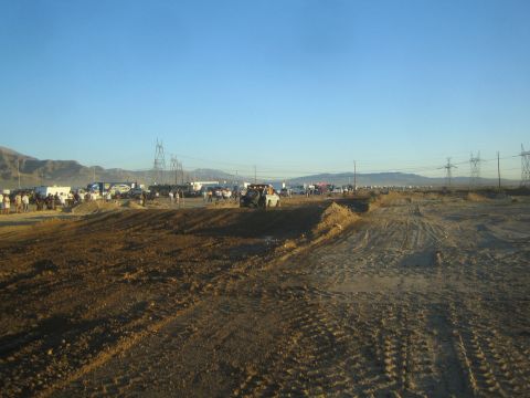 1990 Ford Bronco - churned dirt off-road track stretches across the desert with distant vehicles, spectators and power-line towers under a clear blue sky.