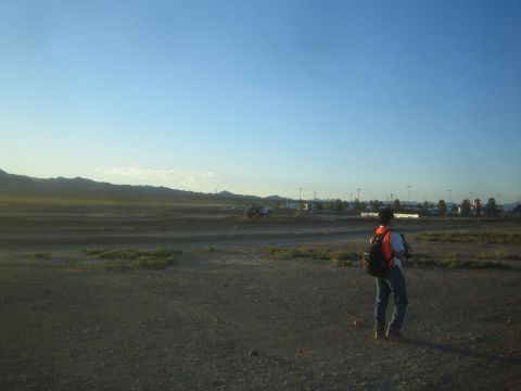 1990 Ford Bronco - person wearing an orange backpack walks across an open desert flat toward a distant off-road course and parking area at dusk.