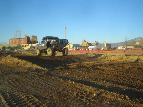 1990 Ford Bronco - lifted off-road pickup truck launches off a dirt berm with a casino, roller coaster and Ferris wheel skyline in the background.