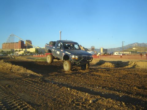 1990 Ford Bronco - black lifted off-road race truck marked 249 catches air over a dirt berm at a racing event with an amusement park skyline behind.