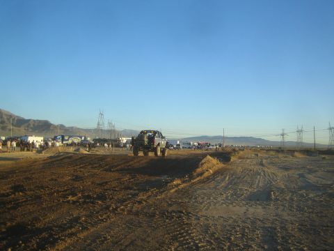 1990 Ford Bronco - off-road truck goes airborne over a dirt jump on a wide desert course backed by transmission towers and parked spectator vehicles.