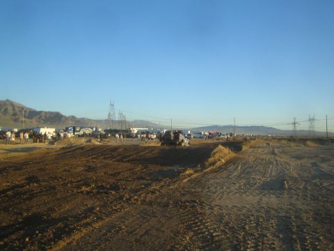 1990 Ford Bronco - broad churned-dirt desert off-road course with distant parked vehicles, spectators and power-line towers under a clear blue sky.