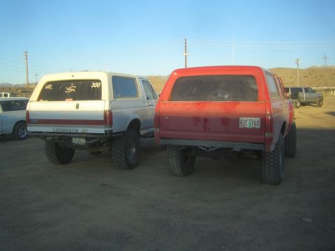 1990 Ford Bronco - Rear view of a white lifted Ford Bronco marked 300 beside a red lifted Bronco with a Colorado plate, parked on dirt at an event.