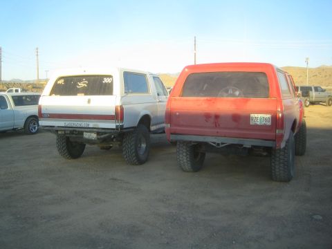1990 Ford Bronco - Rear three-quarter view of a white lifted Ford Bronco marked 300 next to a red lifted Bronco with a Colorado plate on a dirt lot.