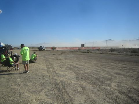 1990 Ford Bronco - Spectators in matching green shirts sit and stand watching a distant truck kick up dust along an orange-fenced desert race course.