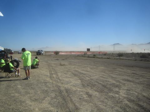 1990 Ford Bronco - group in matching green shirts gathers by camp chairs watching dust rise over an orange-fenced desert off-road course.