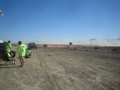 1990 Ford Bronco - Spectators in green shirts watch a truck pass and kick up dust along an orange-fenced desert off-road race course under blue sky.