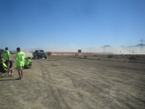 1990 Ford Bronco - off-road truck drives past green-shirted spectators, trailing dust along an orange-fenced desert race course backed by mountains.