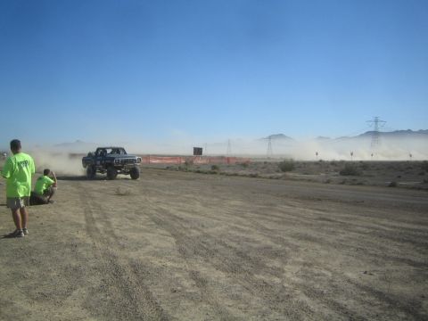 1990 Ford Bronco - off-road truck kicks up dust along a desert race course as a green-shirted spectator watches from the orange-fenced sideline.