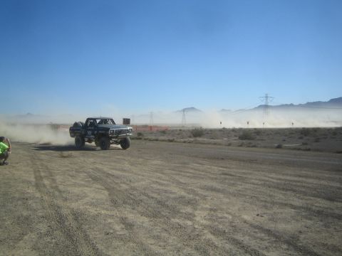 1990 Ford Bronco - dark lifted off-road race truck speeds through clouds of dust along a desert course backed by mountains and power-line towers.