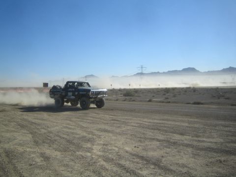 1990 Ford Bronco - dark lifted Ford race truck marked 300 speeds through clouds of dust along a desert course backed by mountains and power lines.