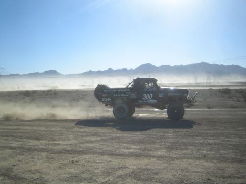 1990 Ford Bronco - black number 300 West Coast Broncos race truck races in profile, throwing up a dust trail under a hazy desert sky.
