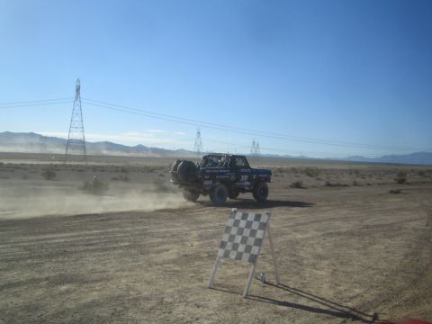 1990 Ford Bronco - black off-road race truck kicks up dust on a desert track with a checkered-pattern course marker standing in the foreground.