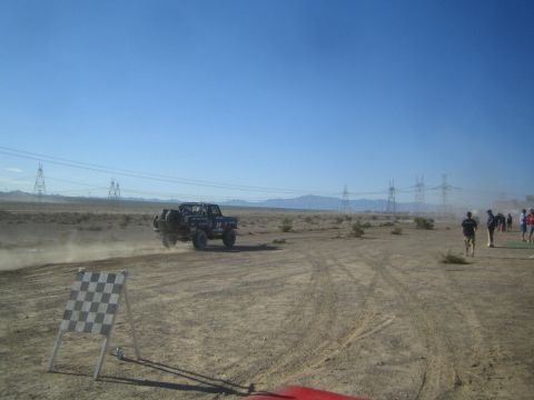 1990 Ford Bronco - dark off-road race truck speeds across a wide desert course as spectators watch from the right beside a checkered marker.