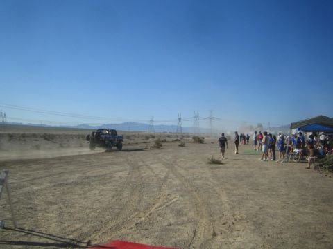 1990 Ford Bronco - dark off-road race truck raises dust on a desert course near a crowd of spectators gathered under shade tents.