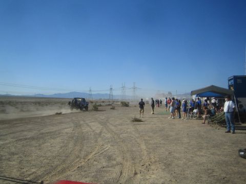 1990 Ford Bronco - Spectators dressed in blue gather under tents along a desert course as a distant off-road race truck trails dust.