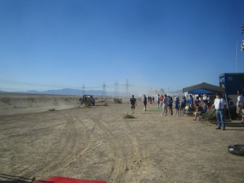 1990 Ford Bronco - crowd of spectators lines a dusty desert race course beside tents and a scoring trailer as a small race truck passes far off.
