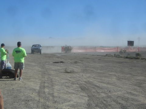 1990 Ford Bronco - blue off-road race truck races along a dusty desert course beside orange fencing while onlookers in green watch from the left.