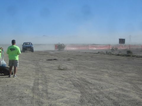 1990 Ford Bronco - blue off-road race truck speeds along a desert course past orange safety fencing while a spectator in green watches.