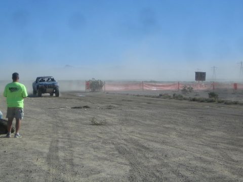 1990 Ford Bronco - blue off-road race truck drives a desert course lined with orange fencing while a green-shirted spectator looks on through the dust.