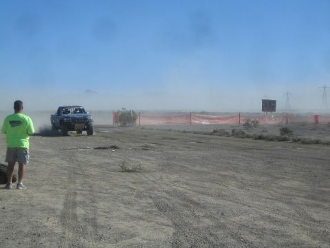 1990 Ford Bronco - blue off-road race truck trails dust along a desert course past orange fencing as a spectator in green stands watching.