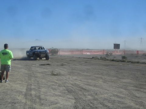 1990 Ford Bronco - blue off-road race truck corners hard through desert dust beside orange course fencing while a spectator watches at left.