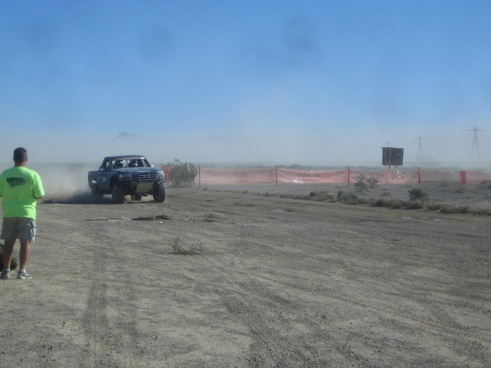 Blue off-road race truck cornering hard in desert dust beside orange fencing