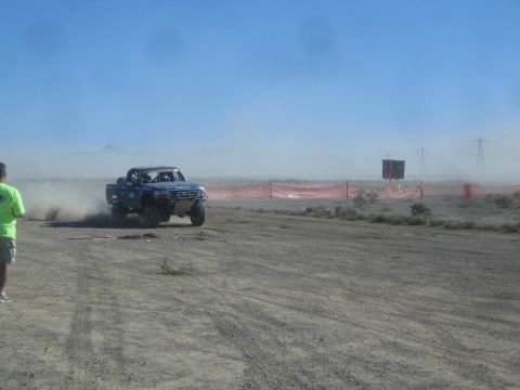 1990 Ford Bronco - blue off-road race truck approaches nearly head-on on a desert course backed by orange fencing and a scoring tower.