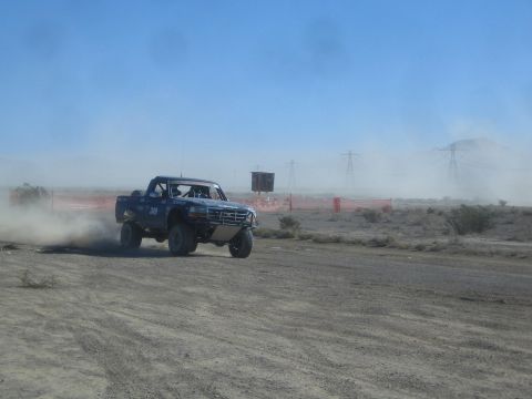 1990 Ford Bronco - blue number 349 off-road race truck powers through swirling dust on a desert course beneath transmission towers.