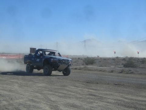 1990 Ford Bronco - blue number 349 off-road race truck races in profile, trailing dust across a desert track with marker flags in the distance.