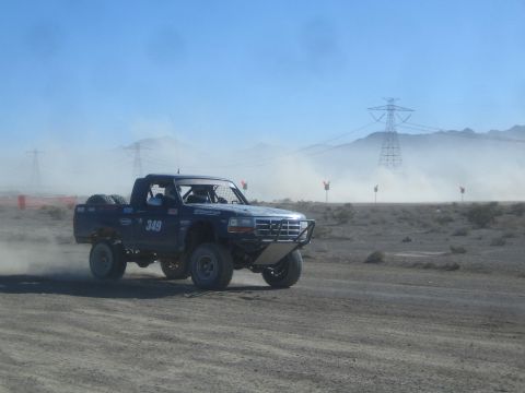 1990 Ford Bronco - blue number 349 off-road race truck drives across open desert terrain backed by a transmission tower and dust haze.