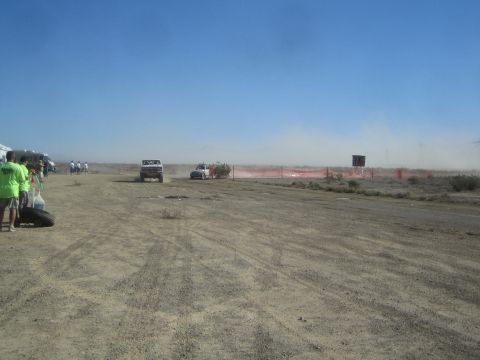 1990 Ford Bronco - Two distant vehicles travel a dusty desert course past orange fencing while spectators stand at the left edge of the track.