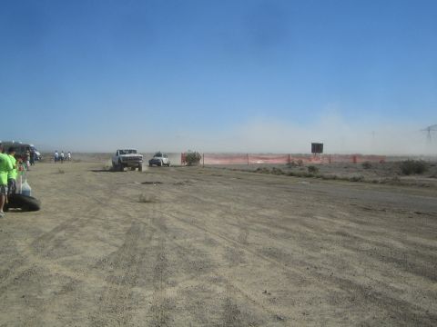 1990 Ford Bronco - light-colored truck and a second vehicle move along a dusty desert course past orange fencing as spectators look on.
