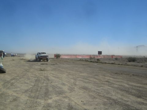 1990 Ford Bronco - white truck trails dust along a desert race course past orange fencing, with spectators at left and a scoring tower beyond.