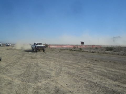 1990 Ford Bronco - white off-road race truck races along a flat desert dirt track, throwing up a long plume of dust near a red course barrier.